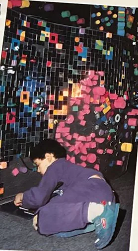 Enfant accroupi posant des modules Piky sur une sculpture d'acier couverte de pièces colorées, 1990